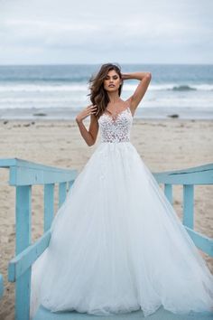 a woman in a white wedding dress standing on a blue railing by the beach with her hands behind her head