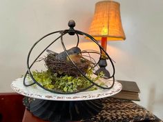 a bird nest sitting on top of a white plate with green plants in it next to a lamp