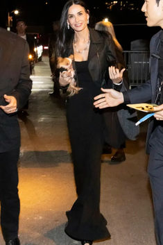 a woman in a long black dress is talking to a man who is holding a small dog