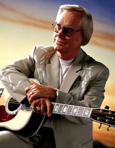 an older man sitting with his guitar in front of a sunset background, wearing sunglasses and a gray suit