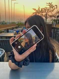 a woman holding up her cell phone to take a selfie with the sun setting in the background