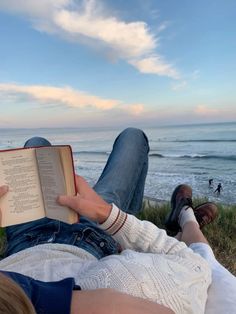 a person laying down reading a book on the grass by the ocean with their feet up