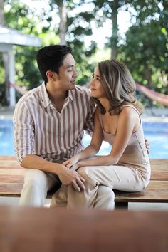 a man and woman sitting next to each other on a wooden bench near a pool