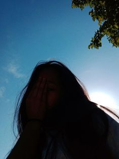 a woman covering her face while standing under a tree with the sun in the background