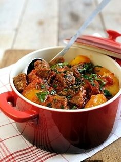 a red pot filled with meat and vegetables on top of a wooden table next to a spoon