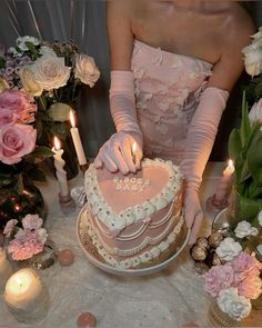 a woman in pink gloves is cutting a heart shaped cake on a table with flowers and candles