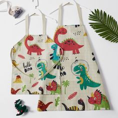 two children's aprons with dinosaurs on them next to a plant and sunglasses