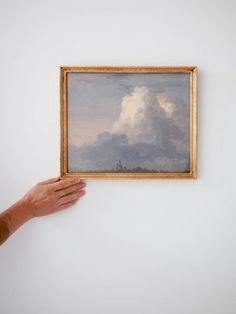 a person holding up a painting in front of a white wall with clouds on it