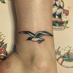 a small bird tattoo on the ankle is shown in black and grey ink, with an old school style ship behind it