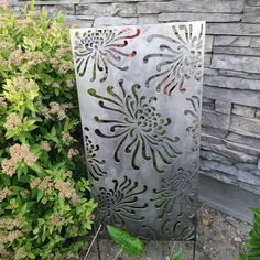 a metal sculpture sitting next to a bush with flowers on it's sides and green plants in the background