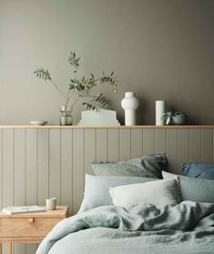 a bed with grey linens and pillows in front of a wooden shelf holding vases