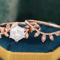 two gold rings with an emerald stone and leaves on the side, sitting on top of a blue velvet cushion