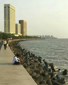 a man sitting on the edge of a pier next to the ocean with tall buildings in the background