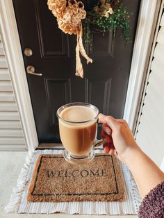 a person holding a cup of coffee in front of a door with welcome mat on it