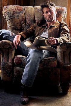 a man sitting in a chair with a cowboy hat on his lap and looking at the camera