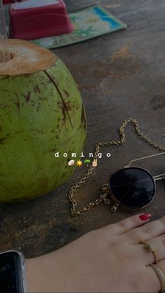 a woman's hand holding a green coconut with sunglasses on it next to a cell phone