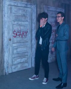 two men standing in front of a door with graffiti on the wall and one man covering his mouth