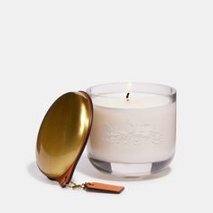 a white candle with a gold lid next to a golden spoon on a white background