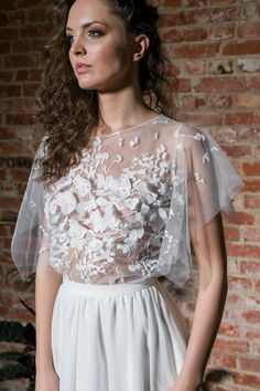 a woman standing in front of a brick wall wearing a white dress with flowers on it