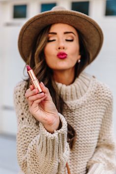 a woman wearing a hat and sweater holding a lipstick in her right hand while looking at the camera