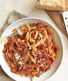 a white bowl filled with pasta and meat on top of a wooden cutting board next to bread