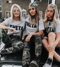 three women sitting on the back of a car wearing matching shirts and camo pants