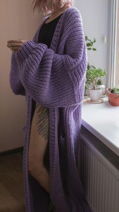 a woman with pink hair is wearing a purple cardigan and standing in front of a window