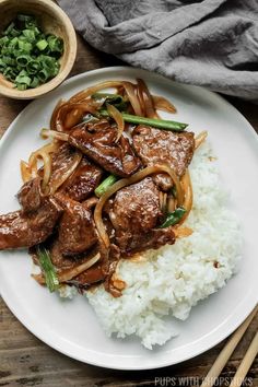 a white plate topped with beef and onions on top of rice next to chopsticks