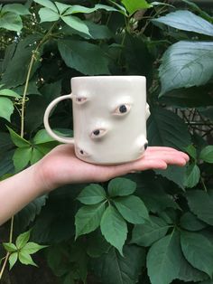 a hand holding a white coffee mug with eyes on it's side and green leaves in the background