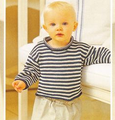 a baby standing next to a white crib and wearing a blue striped shirt with long sleeves