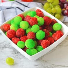 a white bowl filled with green and red gummy balls next to grapes on a marble counter