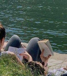 two people laying on the ground reading books next to a body of water with swans in the background