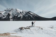 a bride and groom standing on top of a snow covered field with mountains in the background