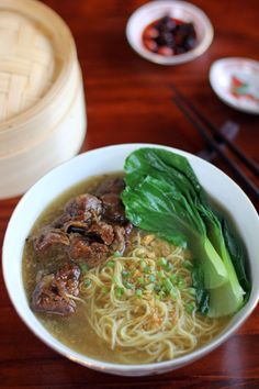 a white bowl filled with noodles and meat on top of a wooden table next to chopsticks