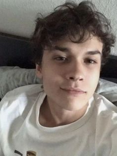 a young man sitting on top of a bed wearing a t - shirt and looking at the camera