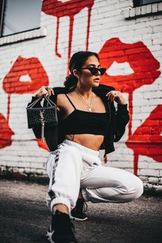 a woman in white sweatpants and black crop top squatting on the ground with her handbag