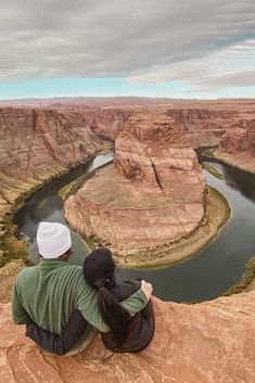 two people sitting on the edge of a cliff looking out over a river and canyon