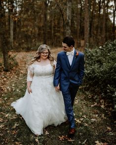 a bride and groom are walking through the woods in their wedding attire, holding hands