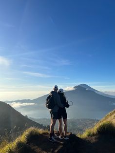 two people standing on top of a mountain looking at the clouds and mountains in the distance