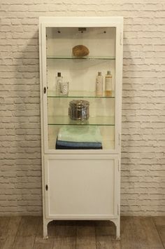 an old white cabinet with glass shelves and bottles on the top, in front of a brick wall