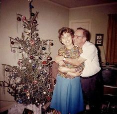 an older man and woman hugging in front of a christmas tree with ornaments on it