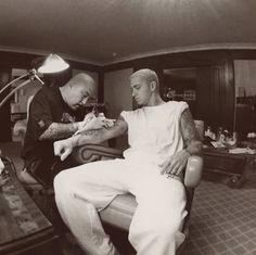 a man getting his arm tattooed by another man in a room with other men sitting around