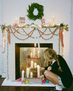 a woman kneeling down in front of a fireplace decorated with candles and garlanded wreaths