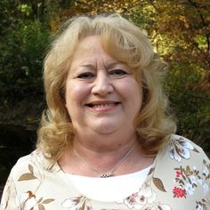 an older woman with blonde hair smiling at the camera while standing in front of some trees