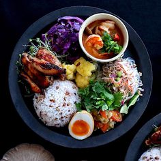 a black plate topped with rice, meat and veggies next to other foods