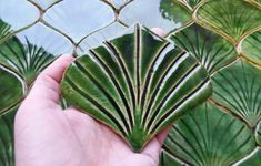 a hand holding a piece of green tile in front of a fence with leaves on it