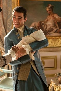 two men in period clothing shaking hands with one holding a doll and smiling at the camera