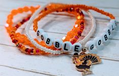 three beaded bracelets with an orange ribbon and a turkey charm on it, sitting on a white wooden surface