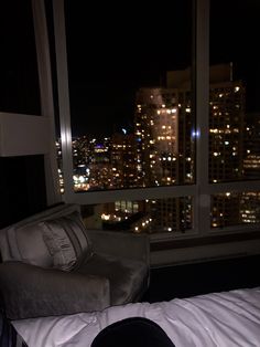 a bed sitting in front of a window next to a night time cityscape