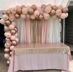 a pink and gold balloon arch is set up on a table with white drapes